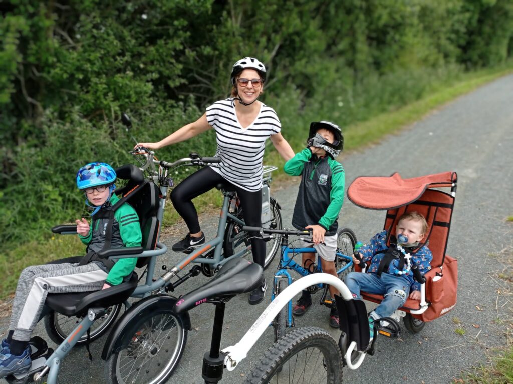 Mother cycles with three children in an accessible bicycle that can cycle with a wheelchair at its front.