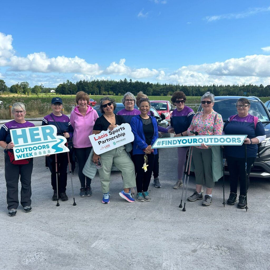 Group of women stand in car park overlooking field holding signage for HER Outdoors Week and #FindYourOutdoors