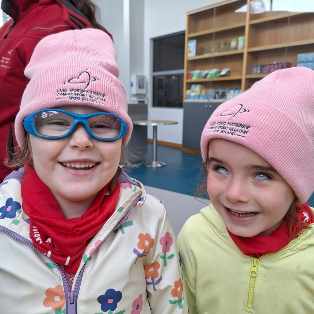 Two young girls smile at camera wearing pink Laois Sports Partnership hats.