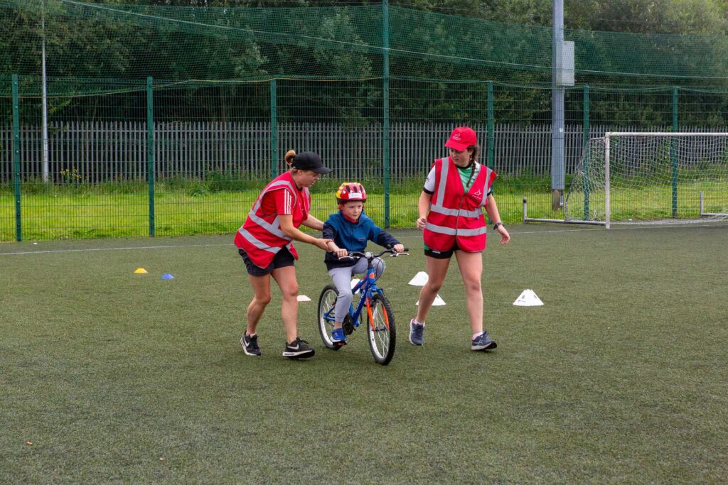 Young boy wearing a bicycle helmet practices cycling with the help of two female instructors.