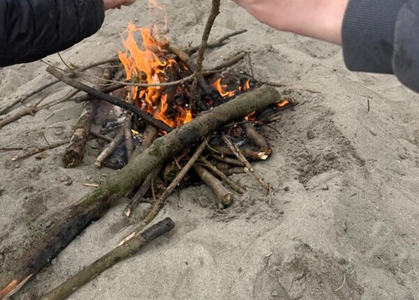 Image of younger adults learning how to create a camp fire, image includes smaller sticks alight.
