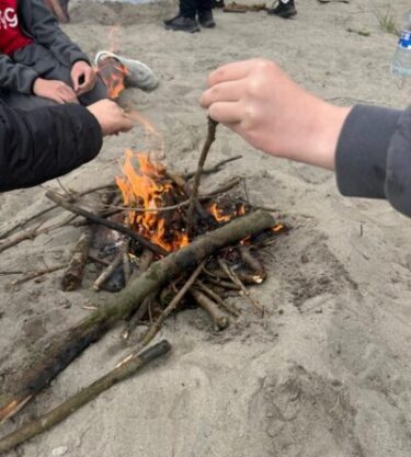 Image of younger adults learning how to create a camp fire, image includes smaller sticks alight.